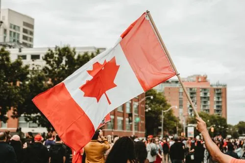 A large Canadian flag is being waved at an outdoor gathering in a city.