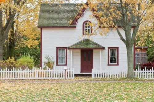 Charming white house with red trim surrounded by autumn trees and a white picket fence.