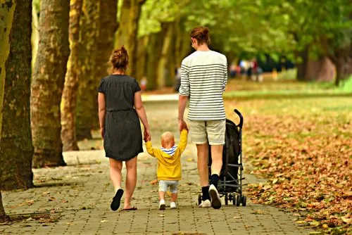 A couple walks with a toddler on a tree-lined pathway, with a stroller beside them.