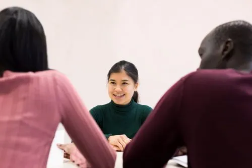Three people are sitting at a table, with a woman in a green sweater smiling at the other two.