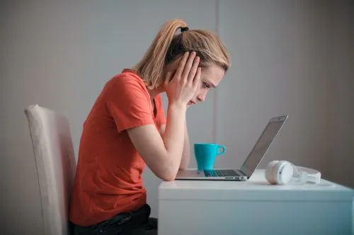 Woman in a red shirt looking stressed while using a laptop at a desk.